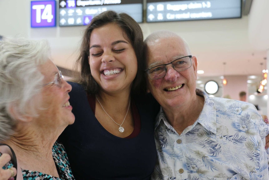A close up of Celeste with her arms around her grandma and grandpa, in an airport terminal with signs in the background.