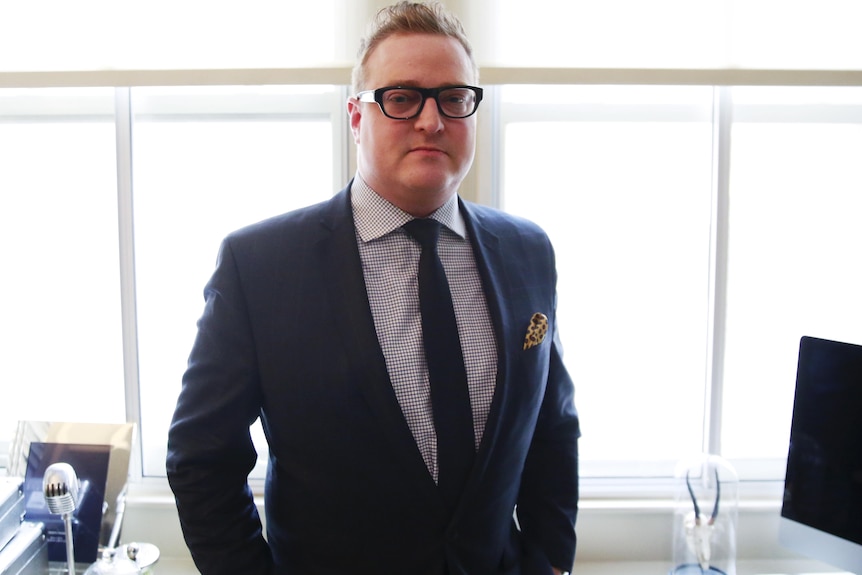 Dylan Howard stands in a suit and glasses in an office.