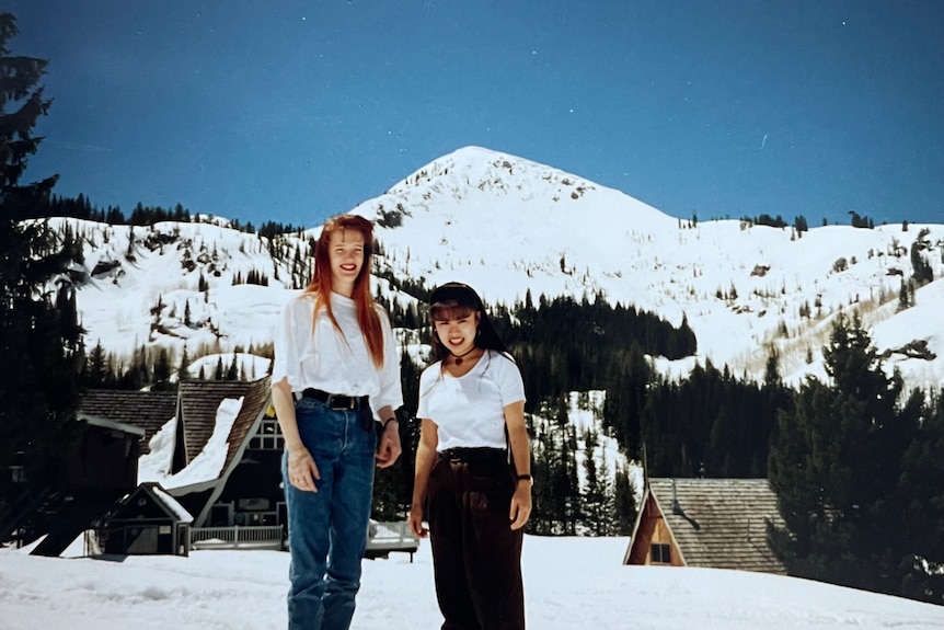 Two women wearing white t-shirts stand outside against a backdrop of snow-covered mountains