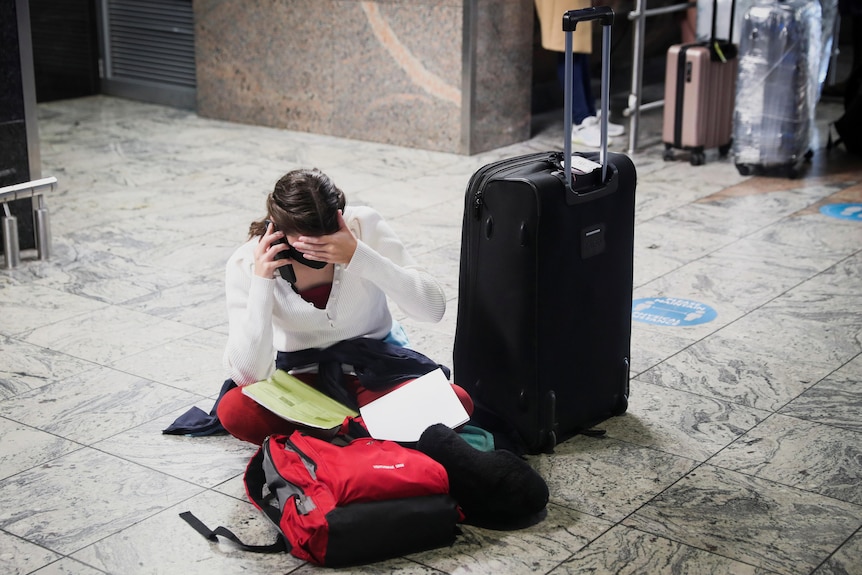 A stranded woman sits on the ground at the airport on the phone with her hand over her face. 