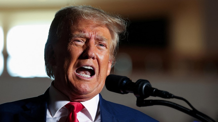 A close-up photo shows an older man with orange-tinged skin speaking loudly into a microphone at a rally.