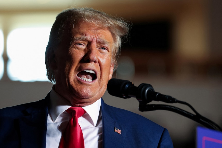 A close-up photo shows an older man with orange-tinged skin speaking loudly into a microphone at a rally.