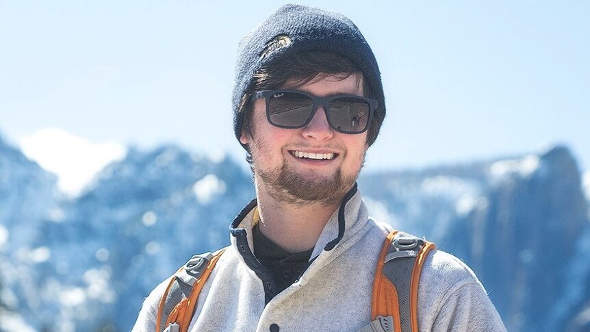 Chase Dekker in a beanie and sunnies holds a camera in front of a mountain scene.