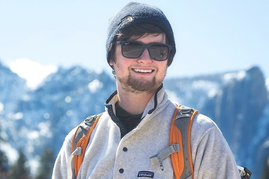 Chase Dekker in a beanie and sunnies holds a camera in front of a mountain scene.