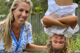 A woman with blonde hair smiling next to her son who's hanging upside down from a swing set with his arms crossed