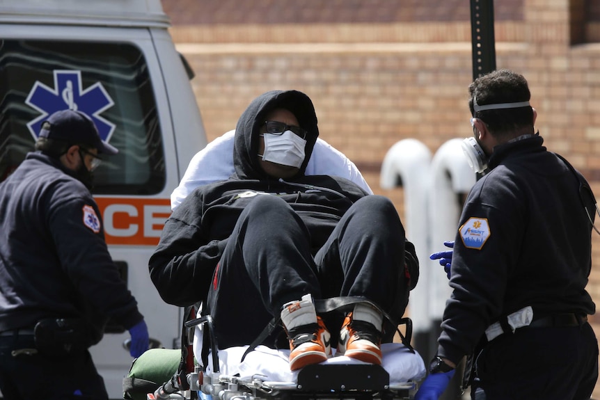 A man in a hoodie and medical mask wheeled from an ambulance by paramedics