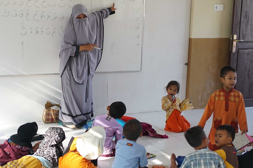 Kids at the Peace Circle learning Arabic. Their teacher is wearing a light purple niqab and points to letters on a white board.