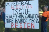 Two men hold a sign that says: "A FEDERAL ELECTION ISSUE FORCED AMALGAMATION ROAD TOLL MIKE BAIRD RESIGN"
