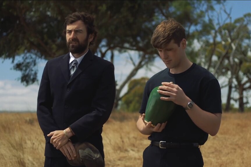 Two men - one in a suit, one in a black t-shirt and pants and a black eye - stand in a dry field with a green urn. 