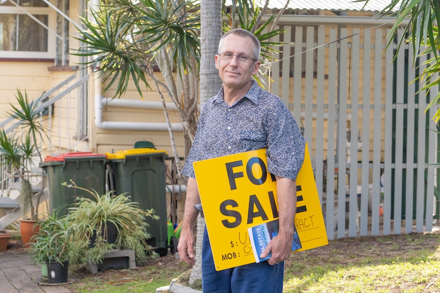 A man stands outside his home with a "for sale" sign and a real estate book under his arm.