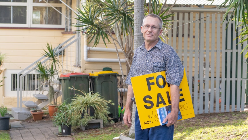 A man stands outside his home with a "for sale" sign and a real estate book under his arm.