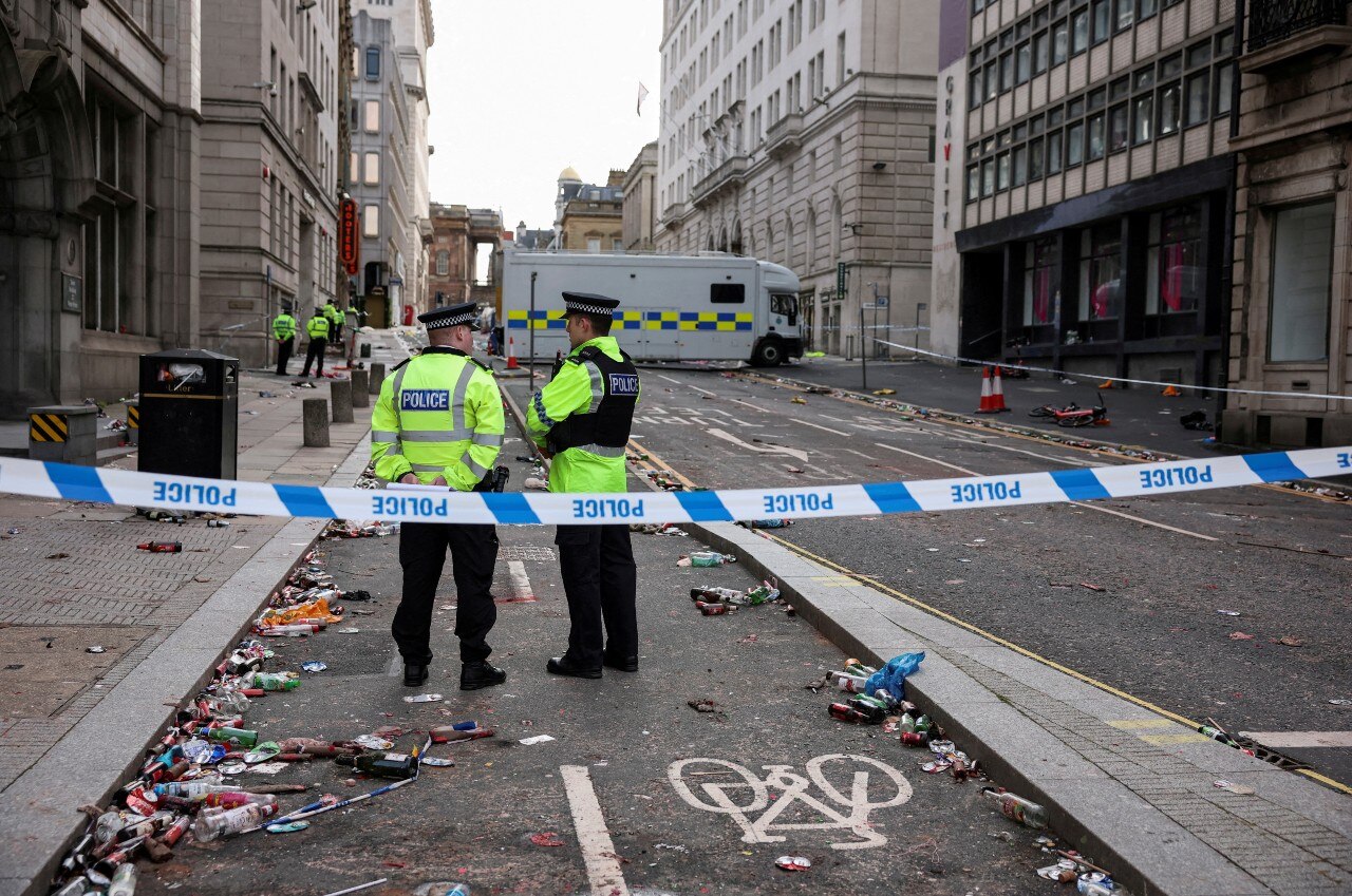Two police officers stand with their backs to the camera on an empty street, which also has been taped off with police tape