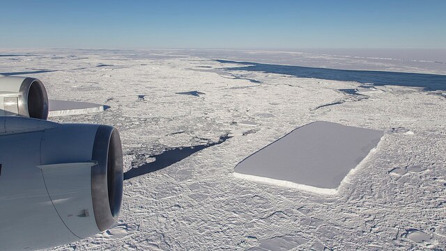 A rectangular-shaped iceberg can be seen in the ocean from the view of an aeroplane, with the berg surround by floating ice.