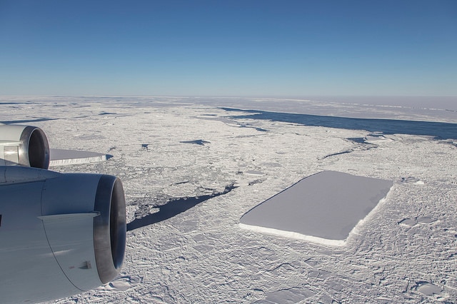 A rectangular-shaped iceberg can be seen in the ocean from the view of an aeroplane, with the berg surround by floating ice.