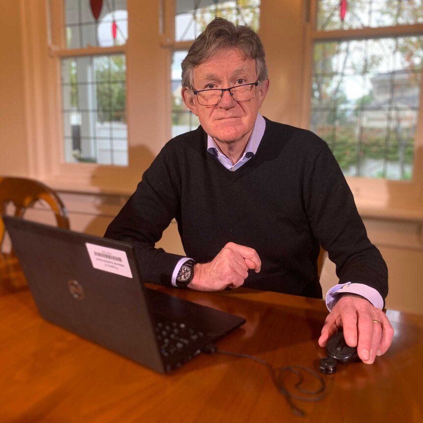 Alan Oster, in glasses, collared shirt and jumper, sits at a wooden dining table with his laptop open in front of him