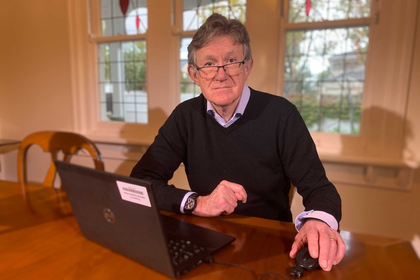 Alan Oster, con gafas, camisa con cuello y jersey, está sentado en una mesa de comedor de madera con su computadora portátil abierta frente a él.