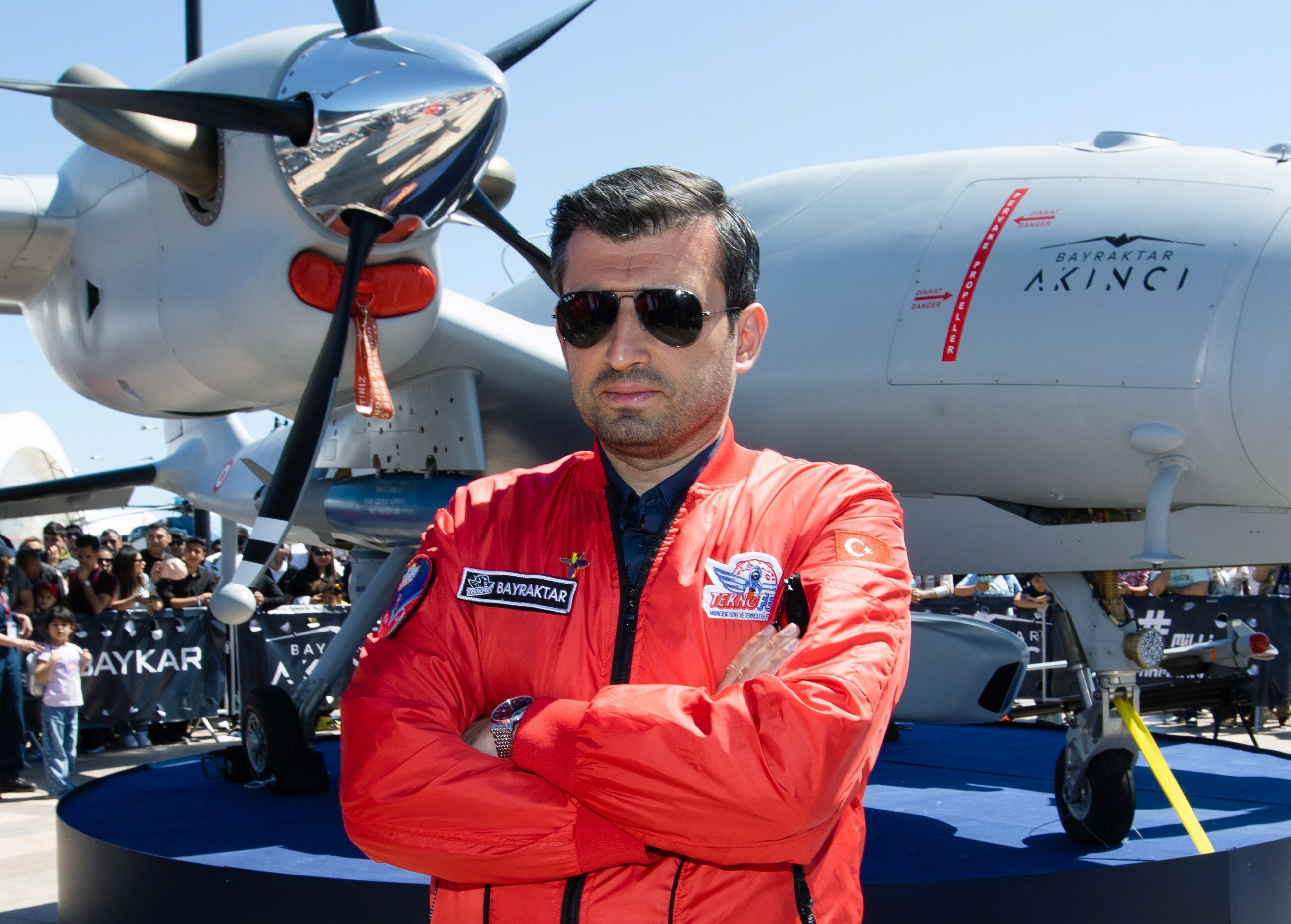 A man in aviator sunnies and a red flight suit folds his arms while standing in front of a military plane
