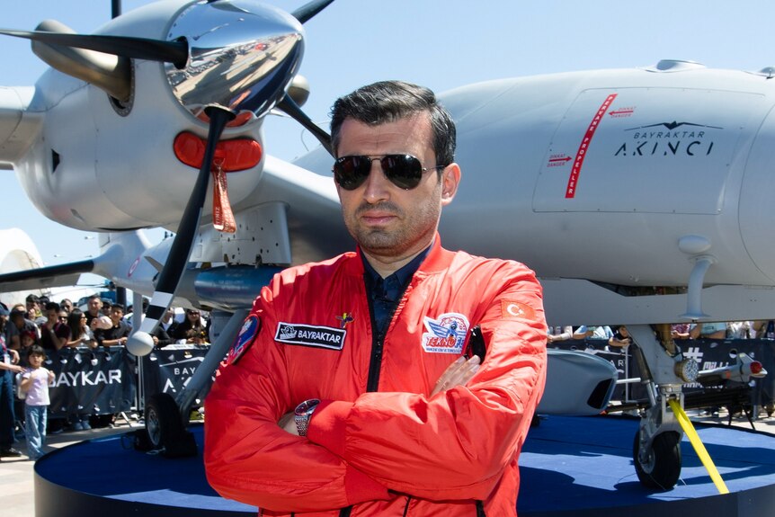 Человек в летном небе и красном летном костюме складывает руки, стоя перед военным самолетом