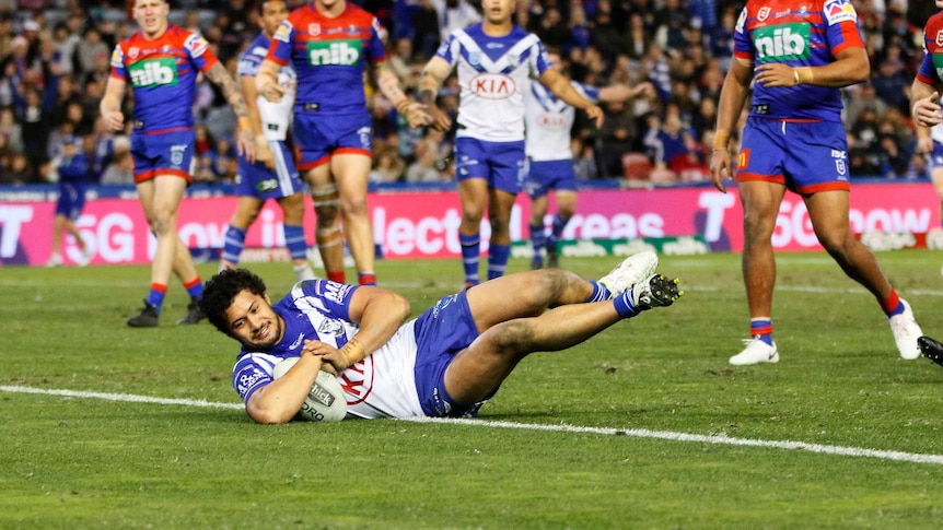 Corey Harawira-Naera lies on his side while grounding the ball. A group of players watch on behind him.