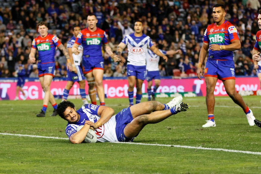 Corey Harawira-Naera lies on his side while grounding the ball. A group of players watch on behind him.