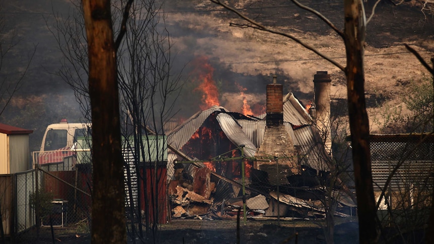 Orange flames break through the melted tin roof a a home destroyed by bushfire. Trees in the foreground are blackened