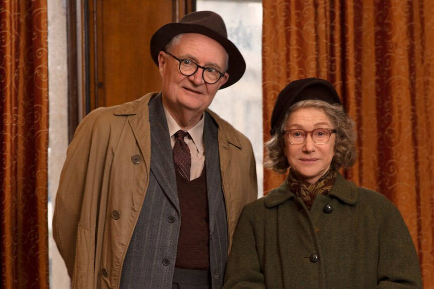 White man and woman in their sixties wear trenchcoats, hats and glasses and look at camera in front of a curtain.
