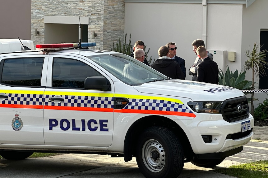 Several men in suits and jackets stand next to a police car outside an off-white suburban house.