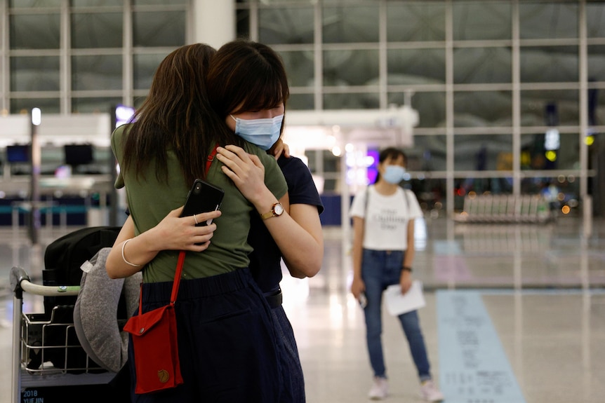 Two women hug in an airport. One's face is visible, she is wearing a mask and closing her eyes as she hugs tight