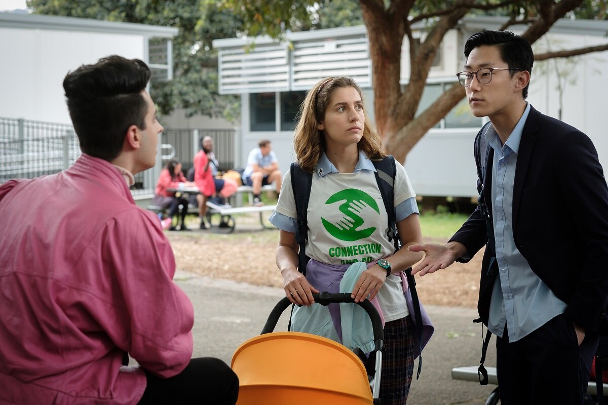 A girl with curly brown hair  wearing white and green shirt with a red pram and man wearing black glasses in a park