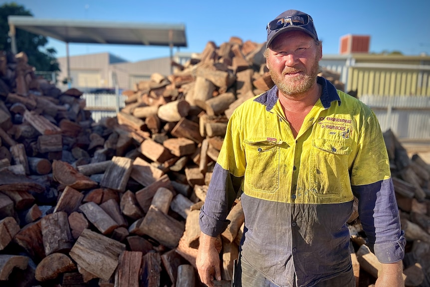 A middle aged man dressed in yellow high-vis with cap,sunglasses & beard stands in front of a large pile of firewood smiling