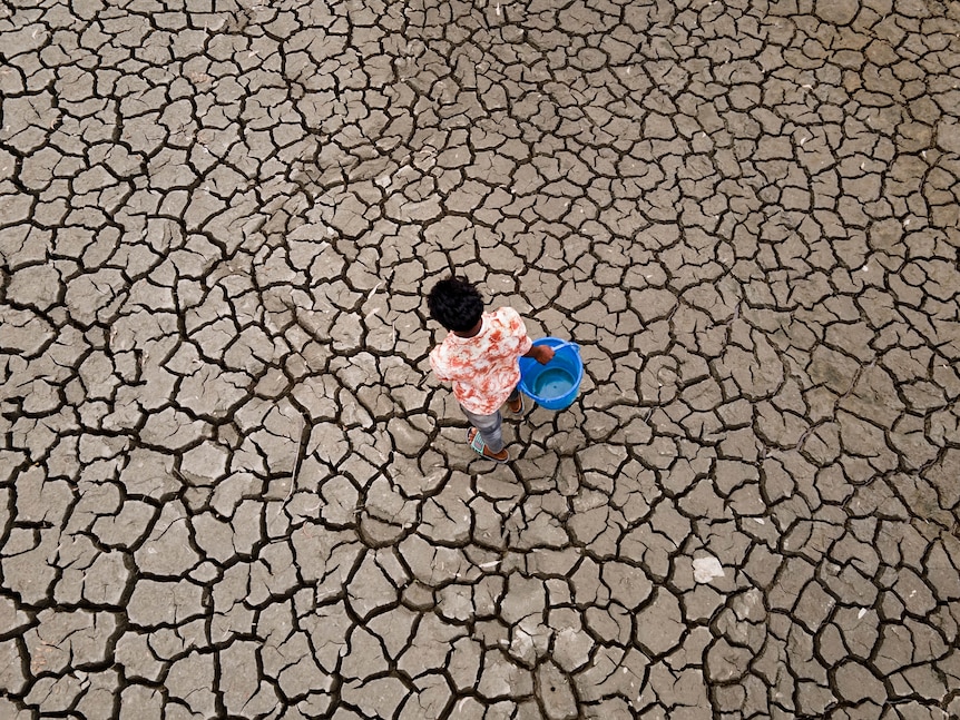 An image from above looking down at a boy carrying a blue bucket, walking across cracked dry land. 