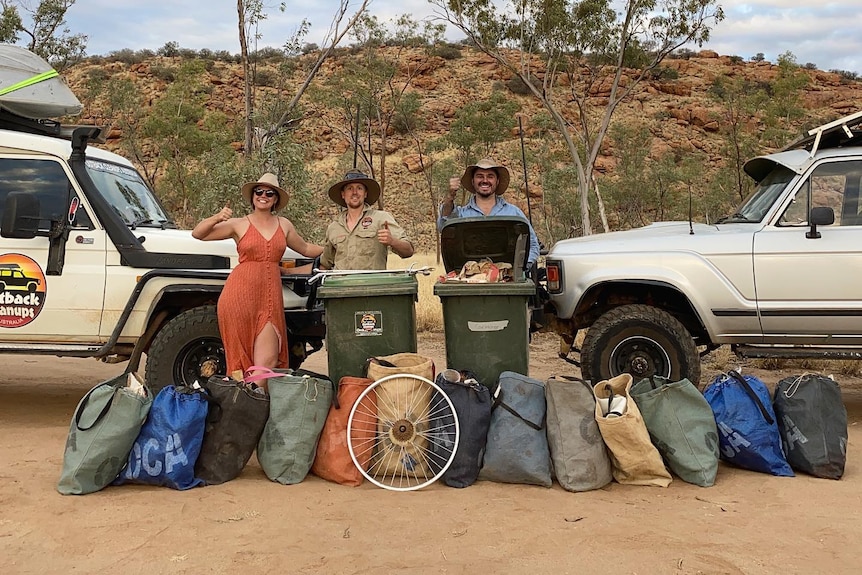 A woman and two men stand with bags of rubbish between two four wheel drives, in front of a rocky outcrop.
