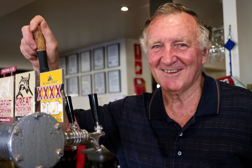 a man smiles at the camera while standing behind a public bar and holding the lever of a beer tap
