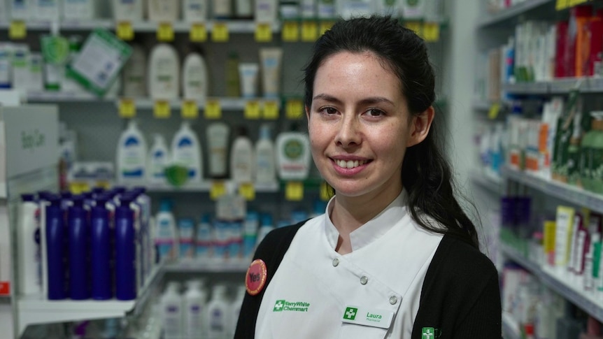 A woman with long dark hair in a pharmacist uniform smiling at the camera inside a chemist.