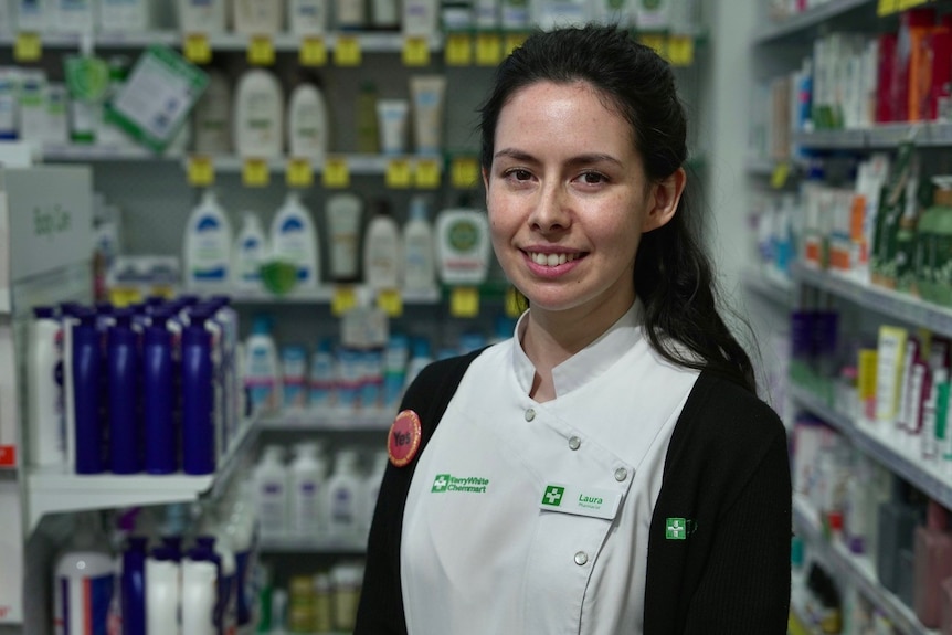 A woman with long dark hair in a pharmacist uniform smiling at the camera inside a chemist.