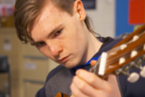 A young boy with short hair makes a face of deep concentration as he plays a classical guitar.