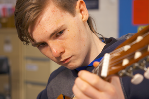 A young boy with short hair makes a face of deep concentration as he plays a classical guitar.