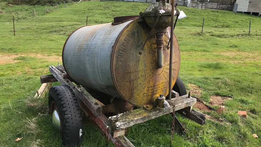A rusted cylinder-shaped metal tank, sitting on a wooden cart in a paddock of green grass.