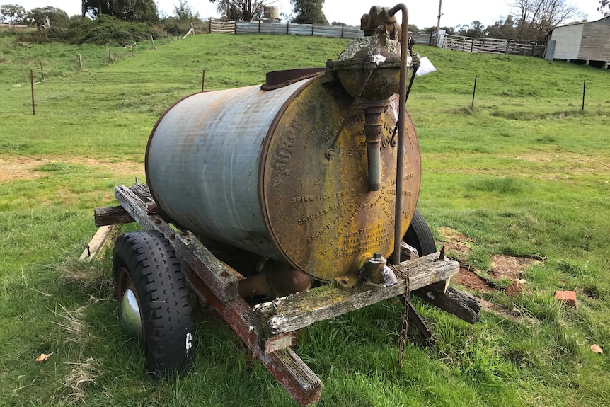 A rusted cylinder-shaped metal tank, sitting on a wooden cart in a paddock of green grass.