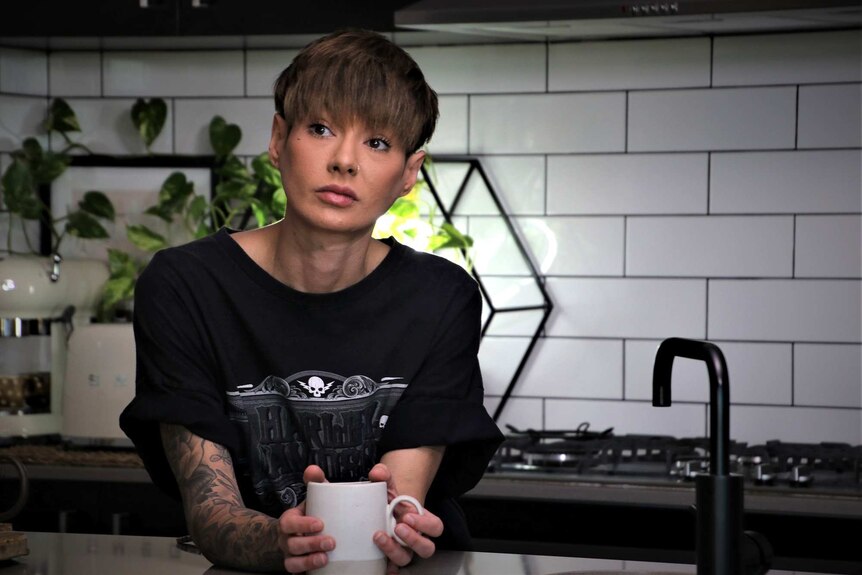 A mid shot of a woman with short hair leaning on a kitchen bench holding a white coffee mug and looking off camera.