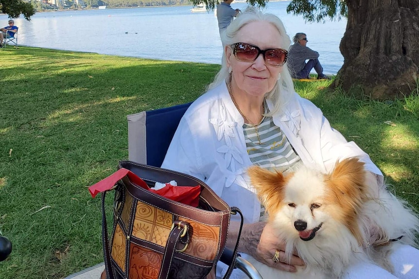 An older lady in sunglasses at the river, holding a dog.
