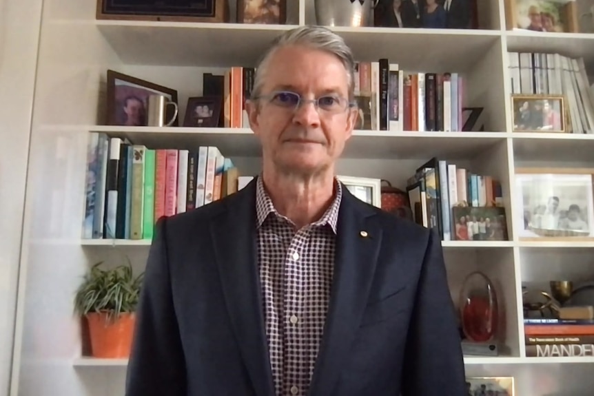 A man in a jacket stands in front a large bookcase.