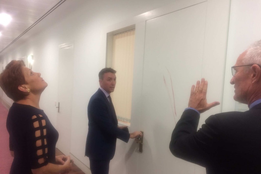 A woman and a two men inspect two vertical lines of blood wiped on a white office door.