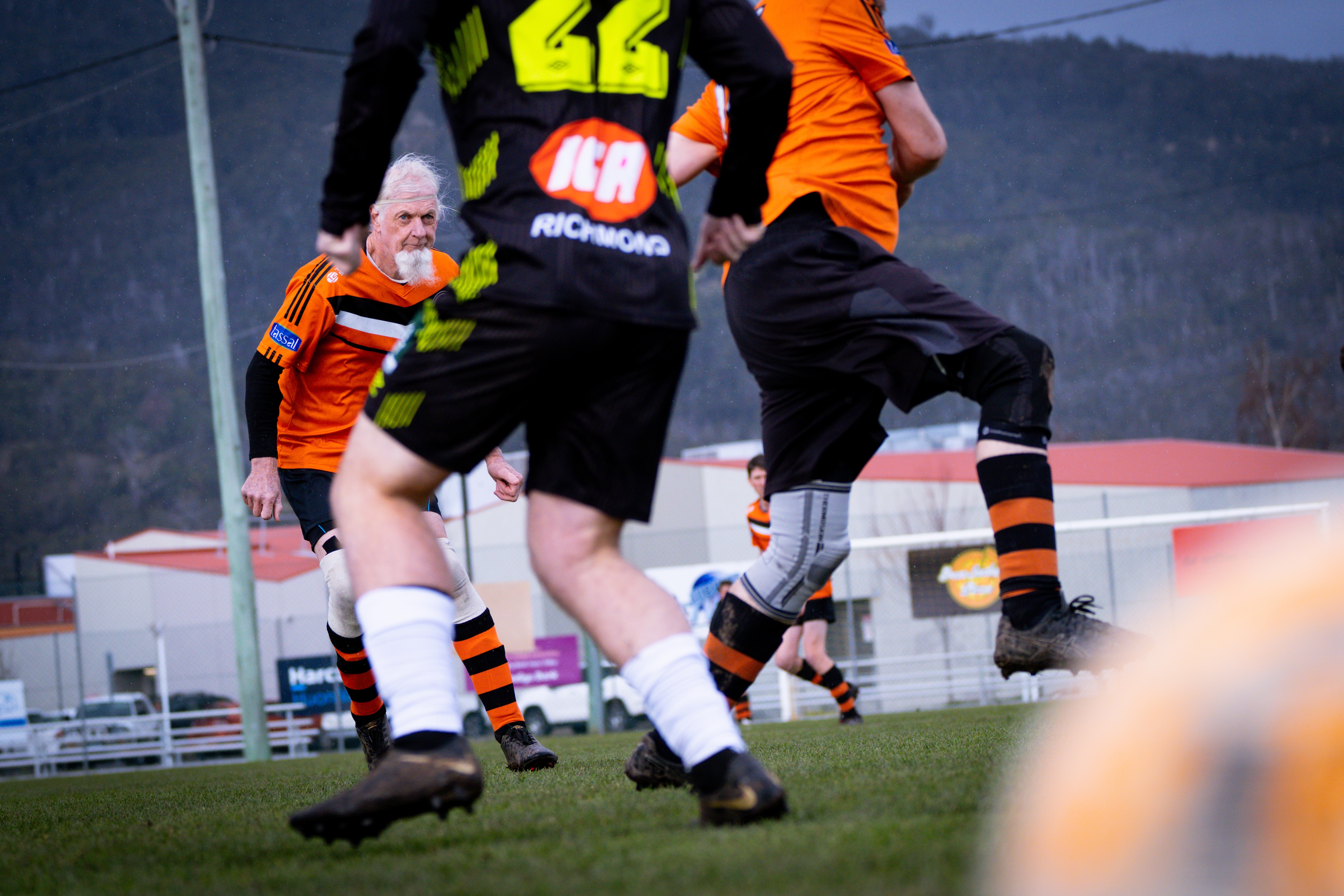 A man with white hair and a long white beard wearing orange soccer colours on a soccer field.