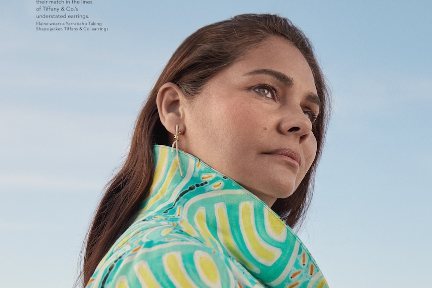 A woman with brown hair and green eyes outside in a green and yellow patterned jumper