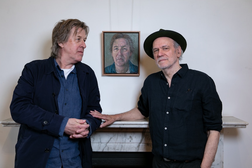 Two middle-aged men stand beside an oil portrait painting of a grey-haired white middle-aged man in a gallery.