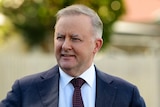 Anthony Albanese looks to the left with a smile on his face. He wears a dark suit with a white shirt and red and blue tie.