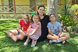 A woman sits on grass with three young children posing for a photo outside a house.