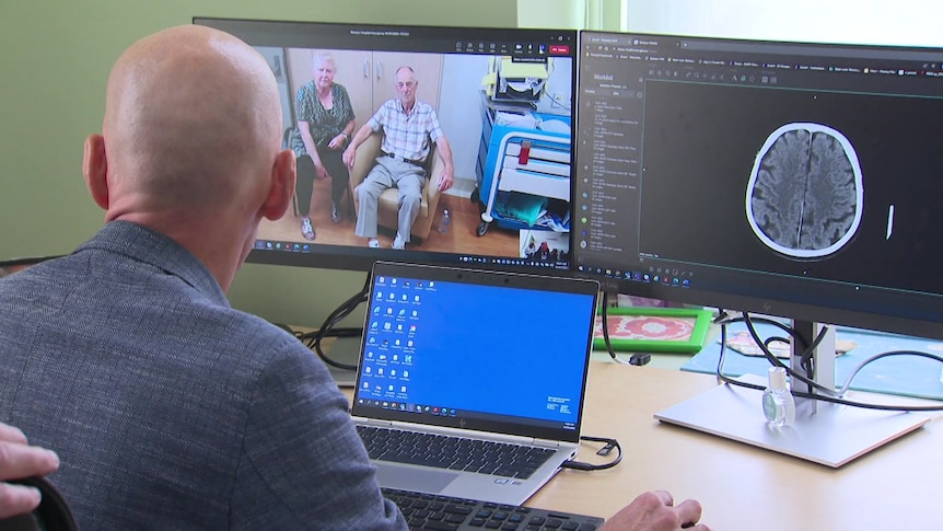 Photo taken from behind a man sitting down and looking at two screens featuring an elderly man and women and a brain scan.