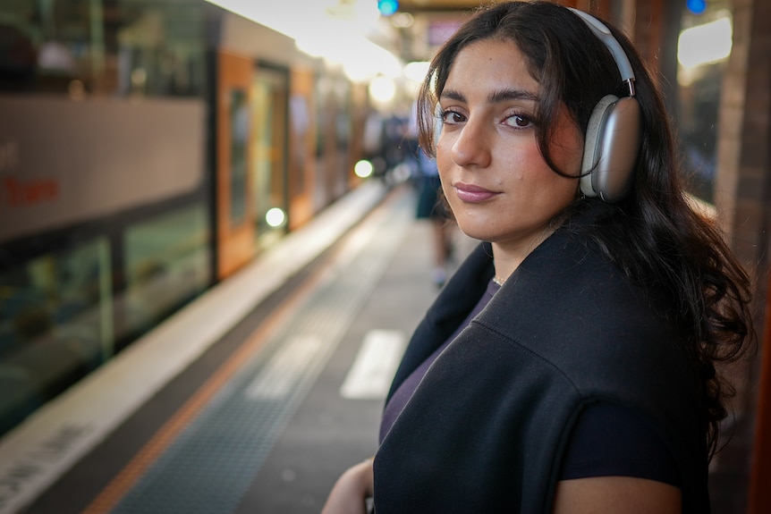 Afsoun Rastegari wearing large headphones and looking into the camera while waiting at a train station.